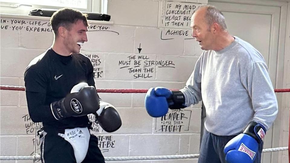 Two men in boxing gloves practising, the younger on the left, an older man on the right