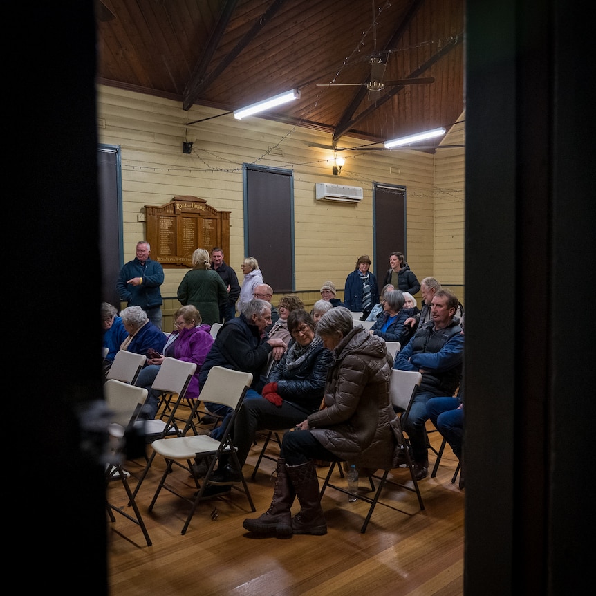 A crowd of people, seen through a doorway, take their seats in a community hall