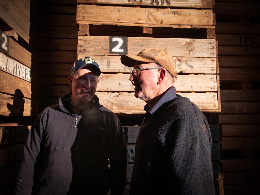 Two men stand next to stacks of wooden crates, sunlight hitting the older man in side profile. They both smile