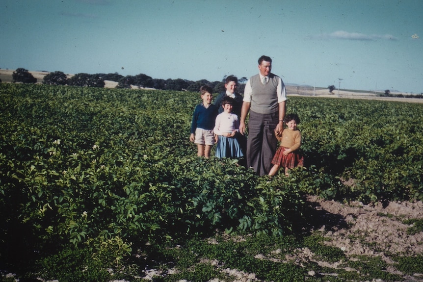 A scanned photo shows a man in grey vest over shirt and tie standing in a field with four children holding hands