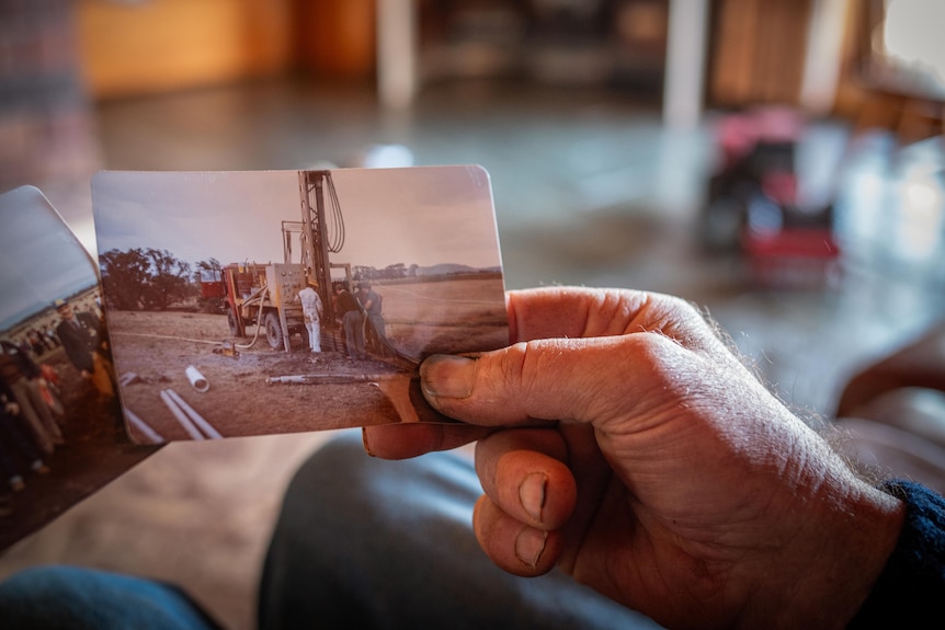 An older man's hand, with dirt under the fingernails, holds a photograph of a younger man standing in front of a tractor