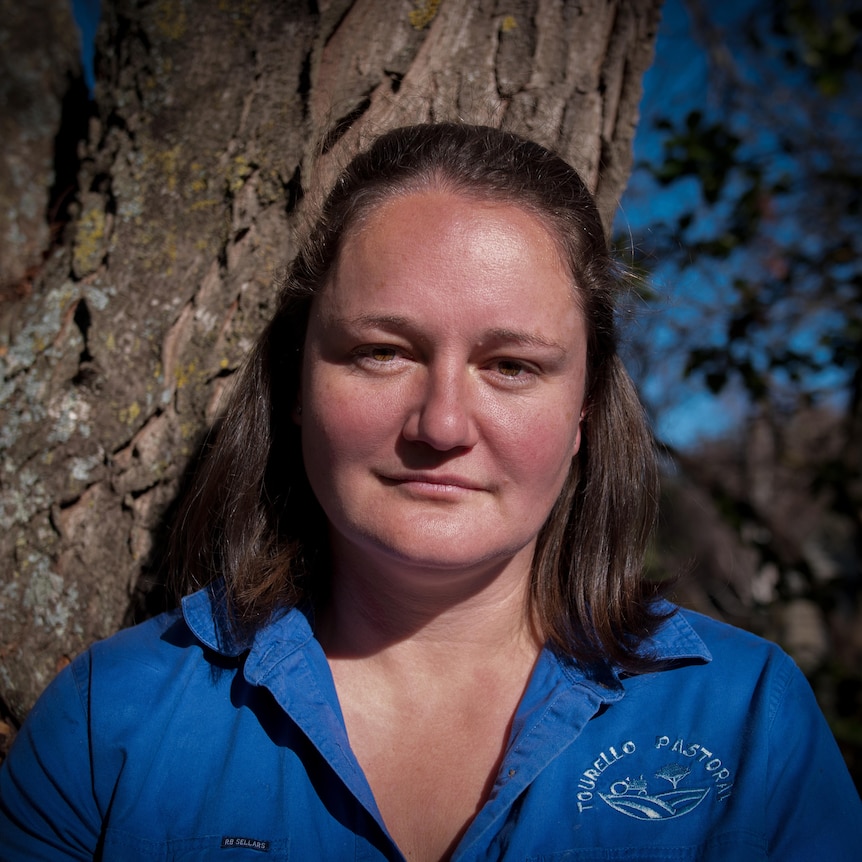 A woman wearing blue shirt stands in front of a tree, looking straight to camera