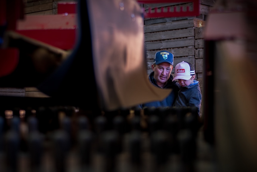 A man holds his young son up to look at machinery inside a shed