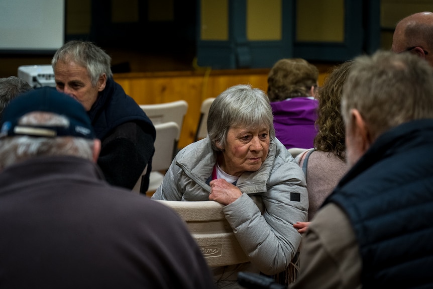 A woman with grey hair, wearing grey puffer jacket, turns around to lean over the back of a chair