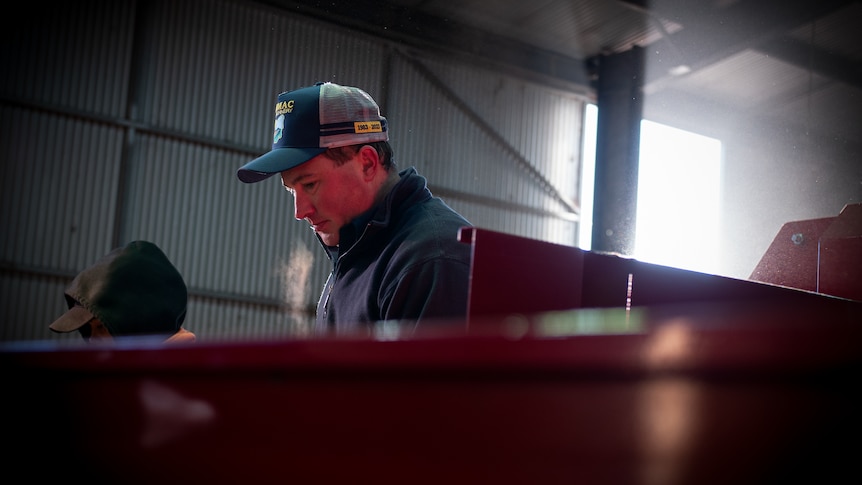 A man in puffer jacket and trucker cap stands behind red machinery. His breath is visible in the cold air