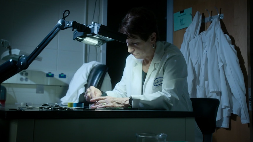 A woman with short haircut wearing a white lab coat looks at a sample on the desk in front of her