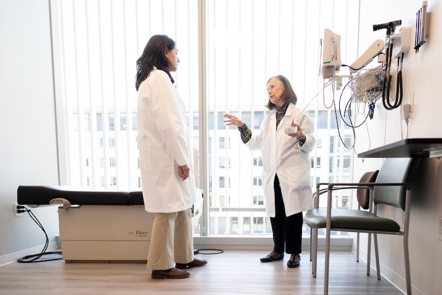 Neurologist Caroline Tanner is talking to another woman in a lab coat in a room of medical equipment