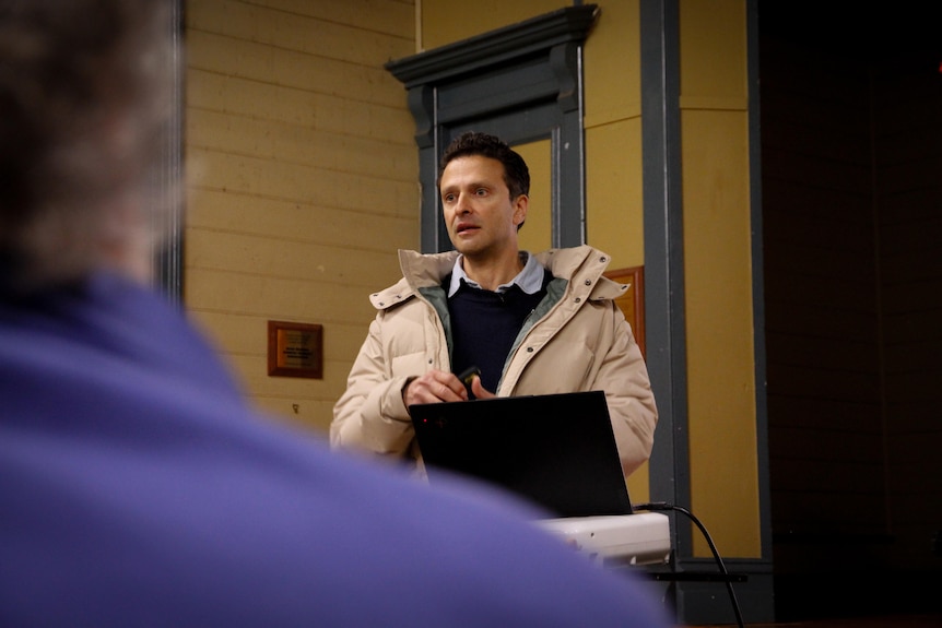 A man wearing dark jumper and beige puffer coat speaks, seen from behind the shoulder of someone wearing purple