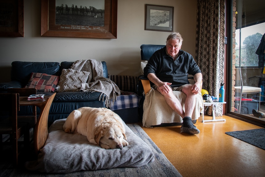 A man sits in a recliner while a golden retriever rests on a large floor cushion, its eyes closed