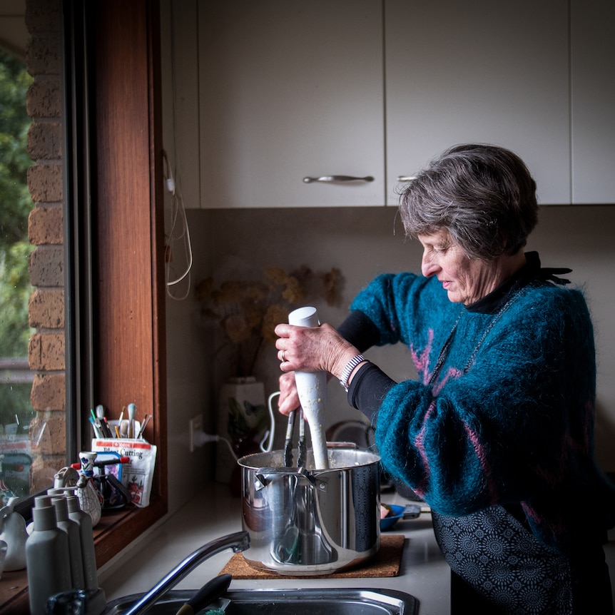 An older woman with dark grey hair stands in a kitchen as she holds an electric mixer in a stainless steel pot