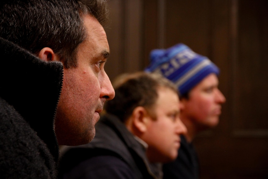 Three men are seen in profile all looking to the same spot, one in focus has a grey collared jumper