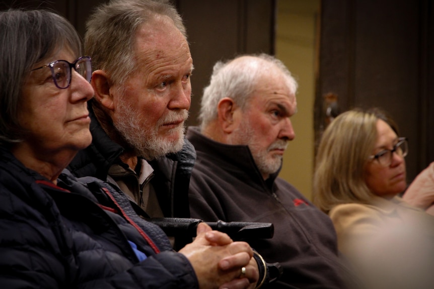 Four people sit side by side with concerned looks on their faces. Two older men are centre of frame