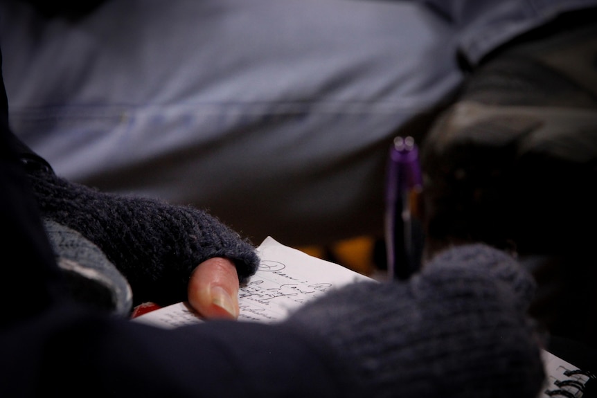 A close-up shows gloved hands holding a pen and notebook, with cursive script in blue ink on the paper