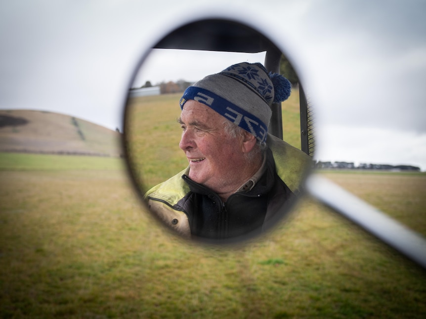 A smiling man in beanie and hi-vis coat is seen in a round wing mirror reflection, against a field of green