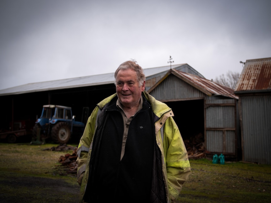 An older man wearing hi-vis jacket over a jumper smiles in front of rusty sheds, on a grey day