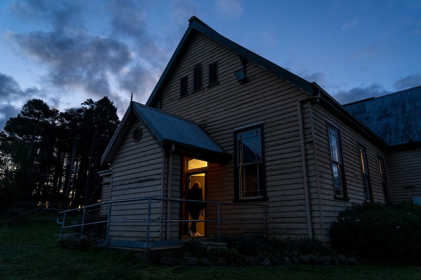 An old community hall, lit from within, is pictured from outside around dusk as the sky turns grey