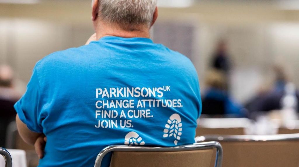 A man where's a Parkinson's UK blue top while sitting on a chair