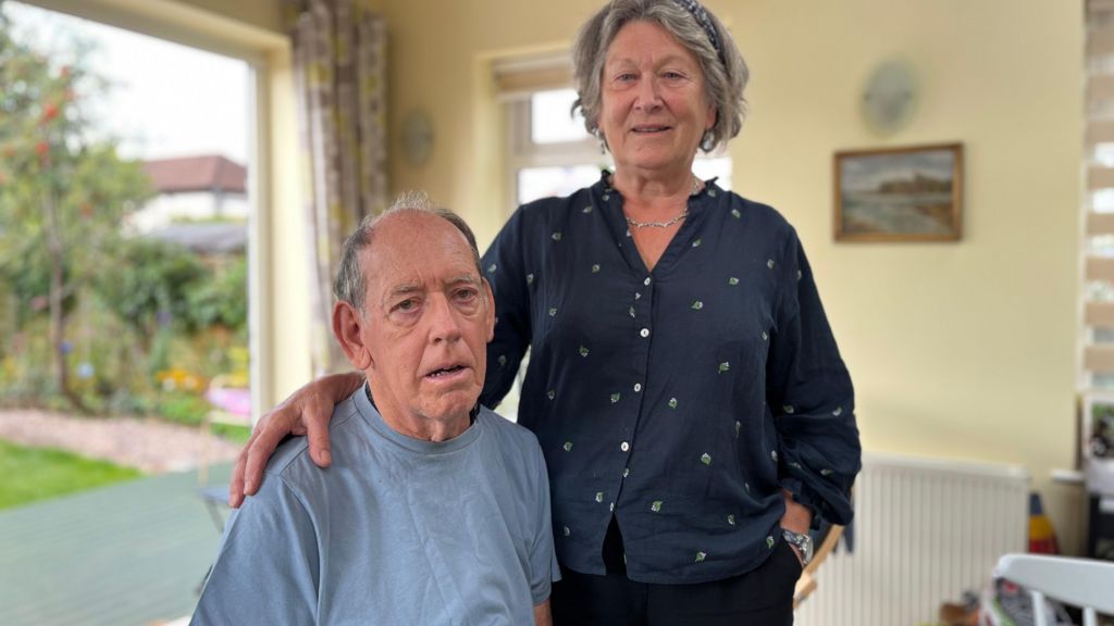 Lynne Telford stands with her husband Rick, who lives with Parkinson's disease, who is sitting down in their home.