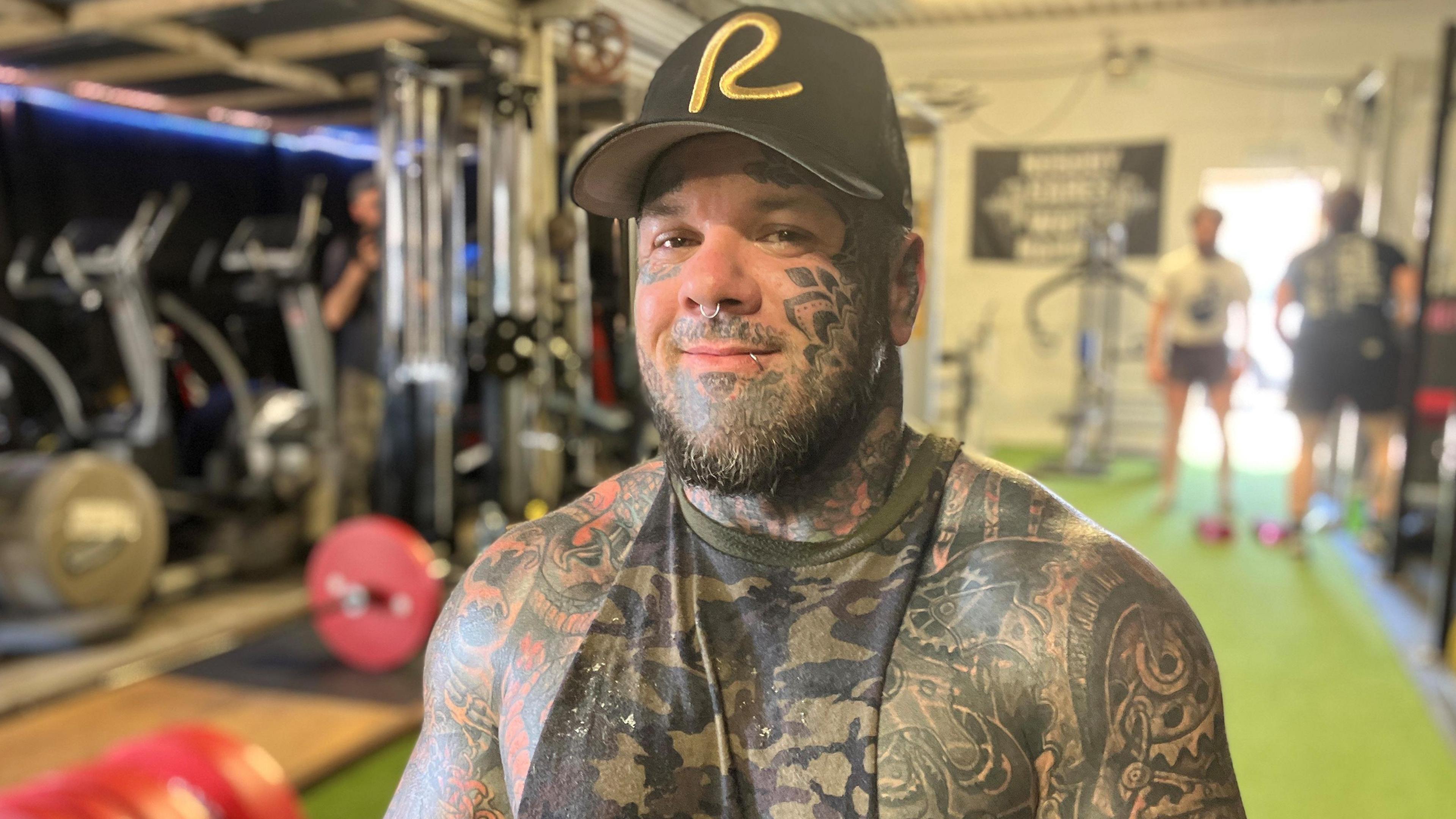 Shaun Slicker in a black cap with a yellow R on it. He is in the gym with weights behind him.