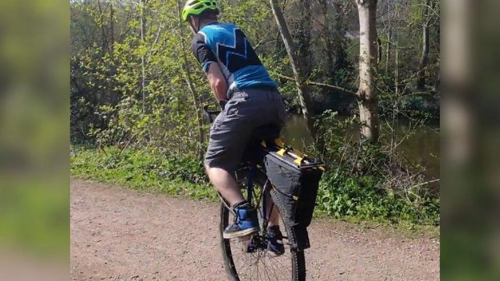 Luke Evison with his back to the camera, on his unicycle, cycling away from the camera. He is wearing his black and yellow cycling helmet and a blue and black cycling t shirt and black long shorts with blue and black trainers. He is on a mud path with trees to the side.