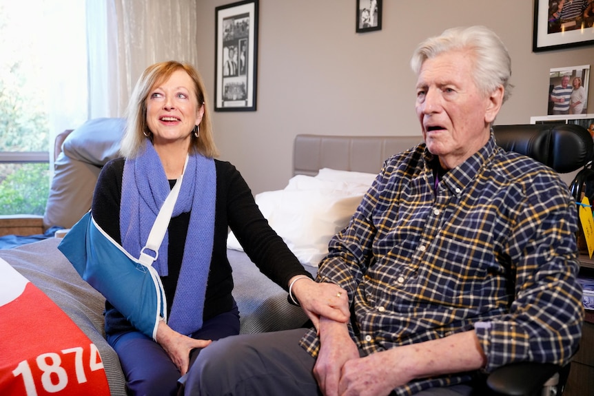 John sits on a couch, a man with grey hair, and Marcia sits beside him, her hand on his.