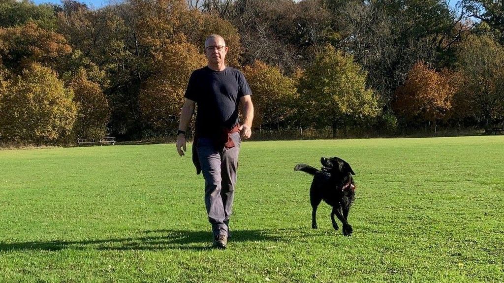 Sam Sharp wearing a dark coloured T-shirt and grey jeans. He is walking through a field with a black dog walking alongside him. 
