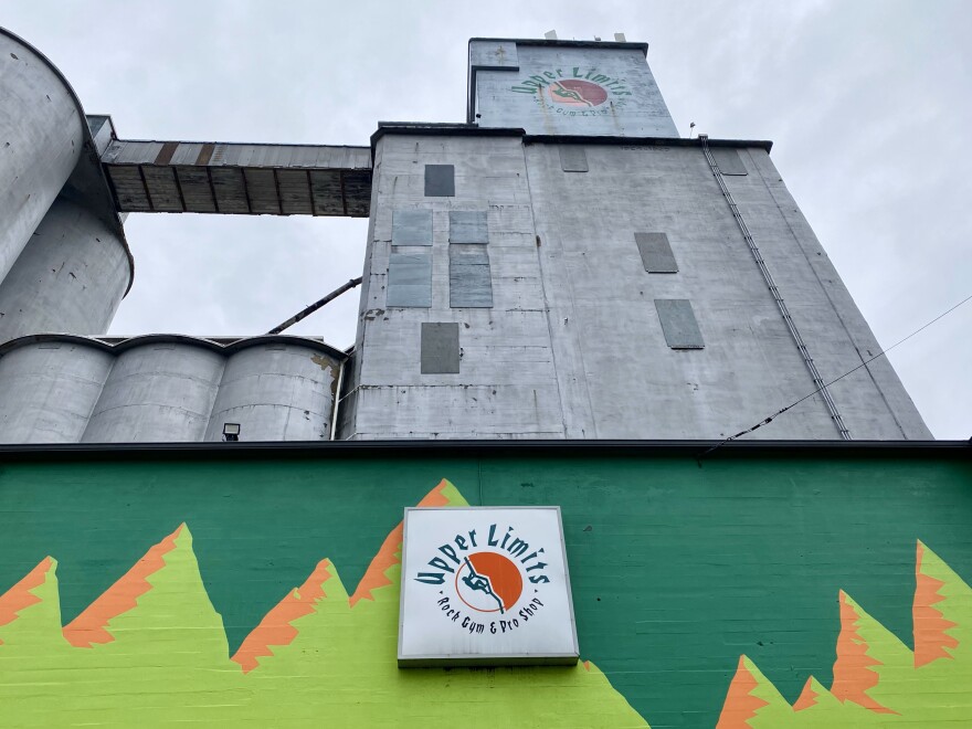 A tall, gray industrial building with a colorful mural at the base featuring mountains and the logo "Upper Limits." The sky is overcast.