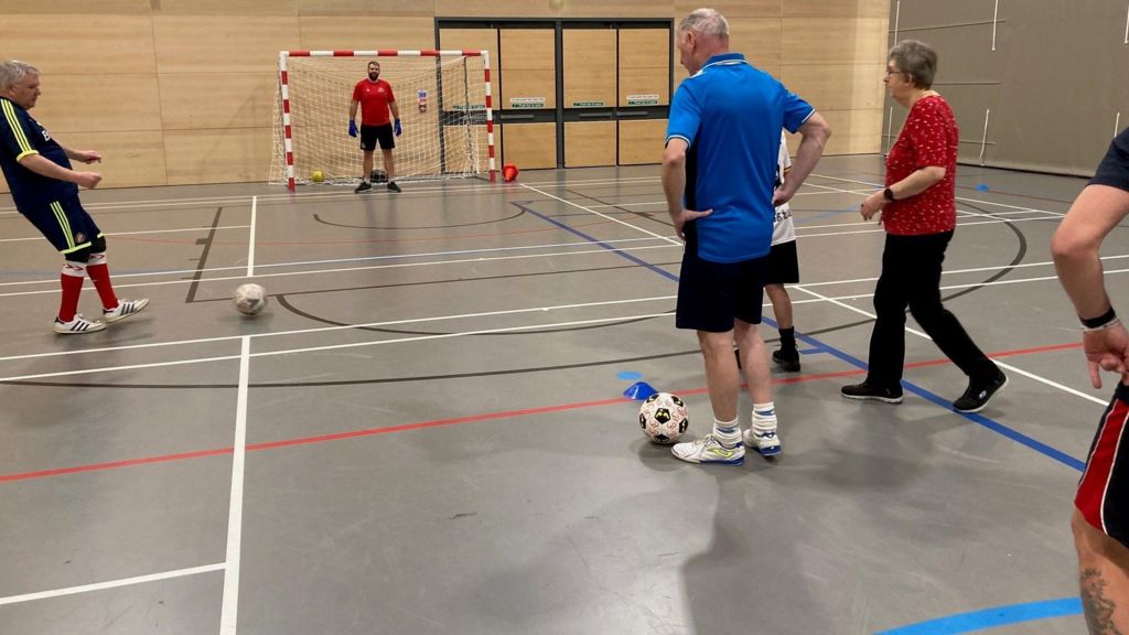 Several people are playing football in a community centre. There is a man standing in the goal and one player is about to kick the ball 