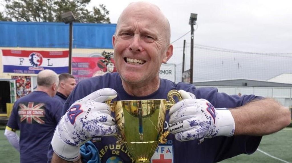 Paul Nicholls holding a gold trophy while smiling. He is stood on an astro turf football pitch while wearing white goalkeeper gloves and a blue T-shirt. It is a grey and cloudy day.