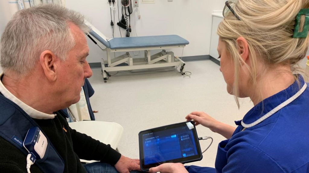 Kevin is with a nurse at hospital as the new system is reprogrammed and switched on. They are both looking at a computer screen, which is wired to his chest. 