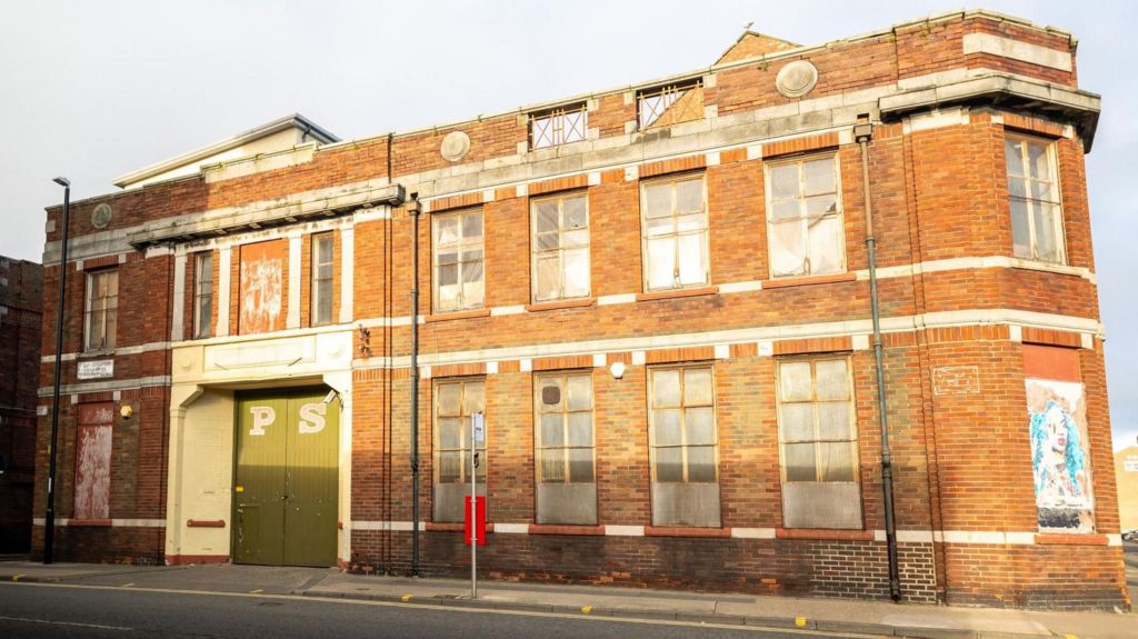 The two-storey red-bricked building has large green doors with the letters "P" and "S" painted on them. There are several large boarded-up windows.