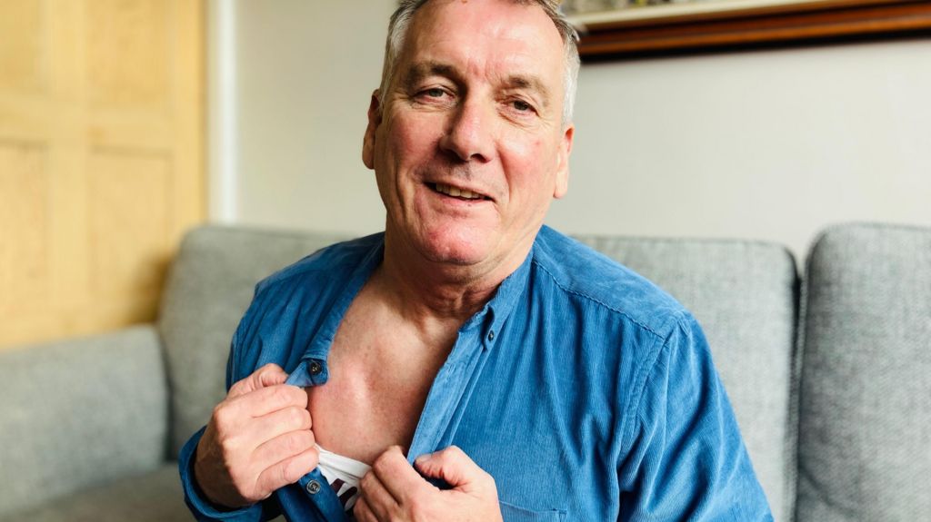 Kevin Hill is sitting on his sofa and has opened his blue shirt to show a lump in his chest. This is where a small computer has been implanted. It is connected to wires that go deep into his brain, to control his Parkinson's disease. 