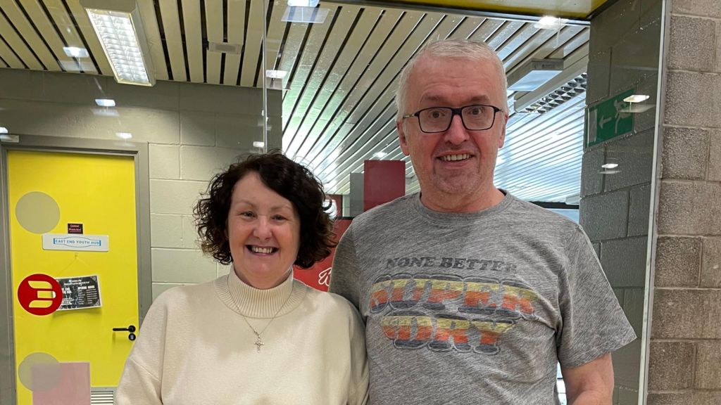 Tony Deeney stands next to his wife, Teresa. Tony is wearing a grey t-shirt and black square glasses to the right of the image. He is smiling while looking at the camera. Teresa has short brown hair wears a cream jumper and stands to the left of the image. She is smiling at the camera.