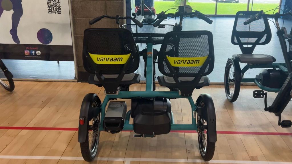 A close up of a side by side tandem tricycle made by Van Raam. There are two seats at the back of the bike with two wheels underneath. There are two handle bars with a basket in between them and one wheel underneath them. Below the double seats you can see the battery for the power assist.