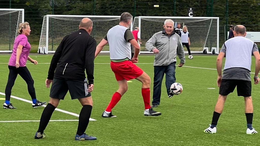 Players with Parkinson's disease taking part in a game at Portishead Town FC. One man with a zipped-up jacket kicks the football.