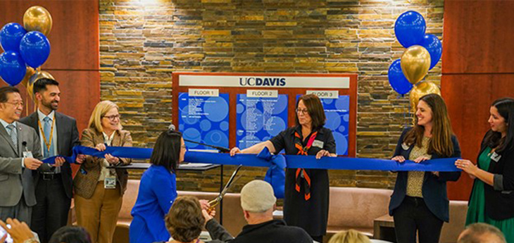Six people are holding a blue ribbon as Malhado-Chang is trying to cut it with a large pair of scissors. Blue and gold balloons in the background.