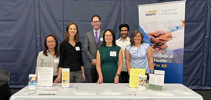 Six persons are standing behind a table with a white cloth, written on it UC Davis Health, Department of Neurology. Everyone smiling to the camera.