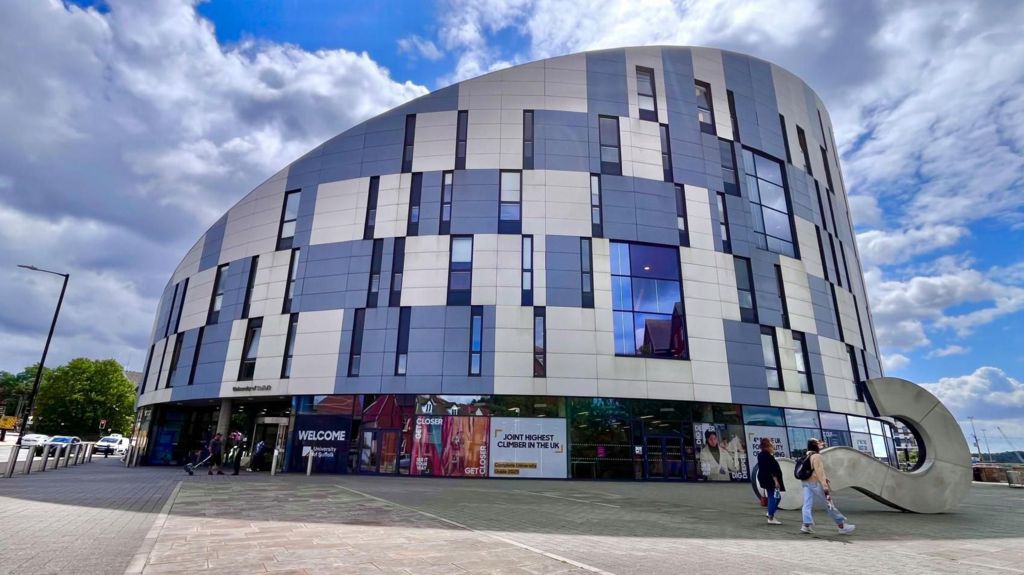 The University of Suffolk building. It is a large sloped building covered in windows and blue and white panels. Two people are walking past with others entering one of the doors. A large question mark sculpture is lying down on the right of the image. A blue sky with white clouds is overhead.