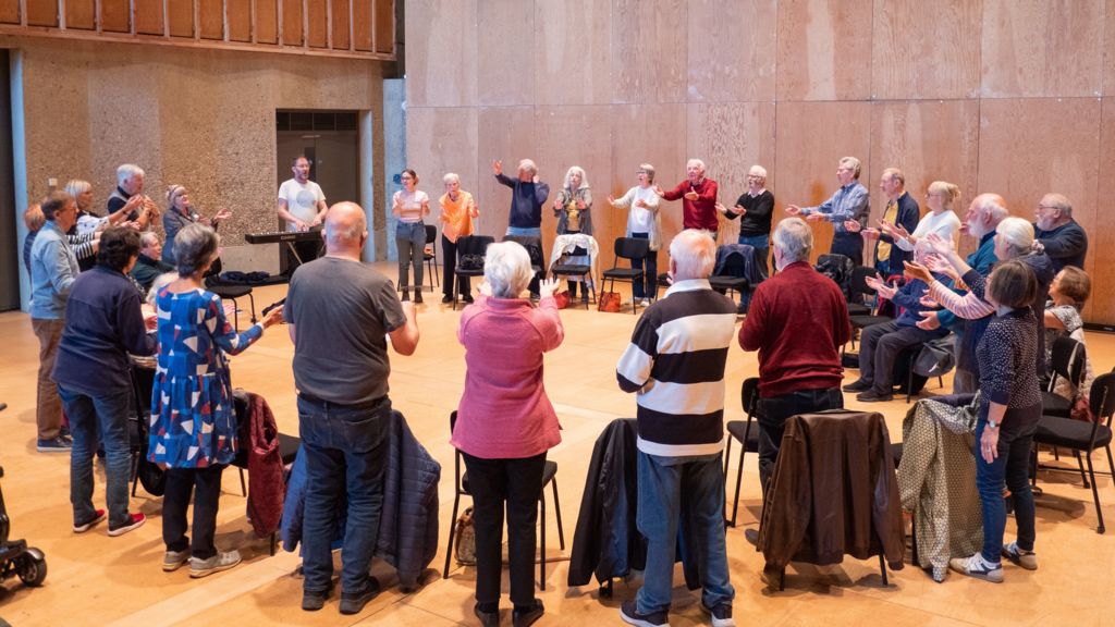 A large group of people stand in a circle, behind their chairs, as they sing. They have their hands lifted in front of them as they sing. They are within a large hall.