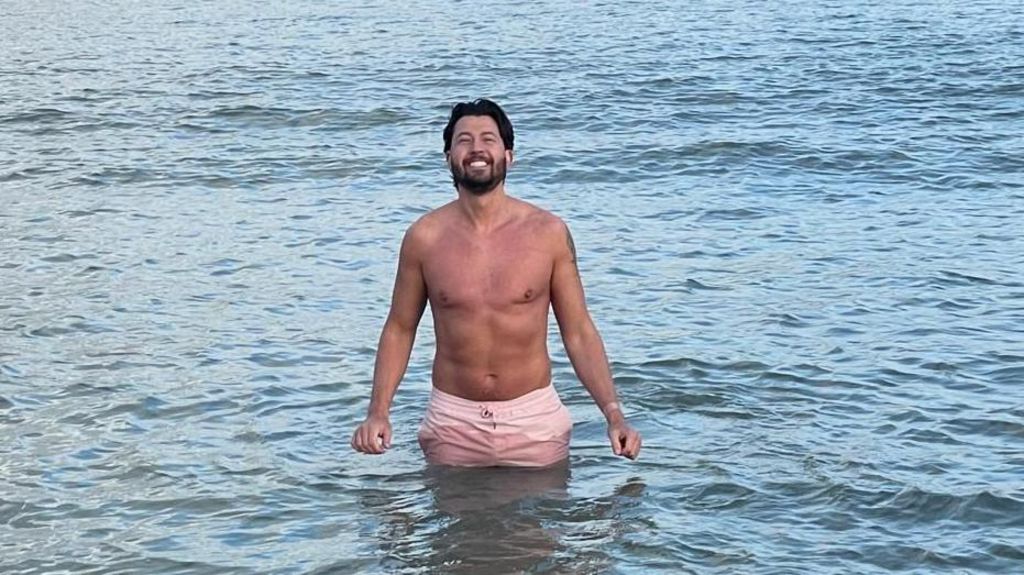 James Madeley is wearing pink swimming trunks, he has brown hair and a brown beard. He is stood in the sea.