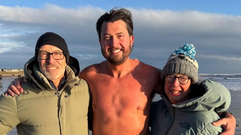 Steven Madeley is wearing a black hat, glasses and a green jacket. He has a white beard. James Madeley is wearing light swimming trunks, he has brown hair and a brown beard. Carol Madeley has a grey, white and blue bobble hat, glasses and a grey jacket. They are all smiling and stood in front of the sea.