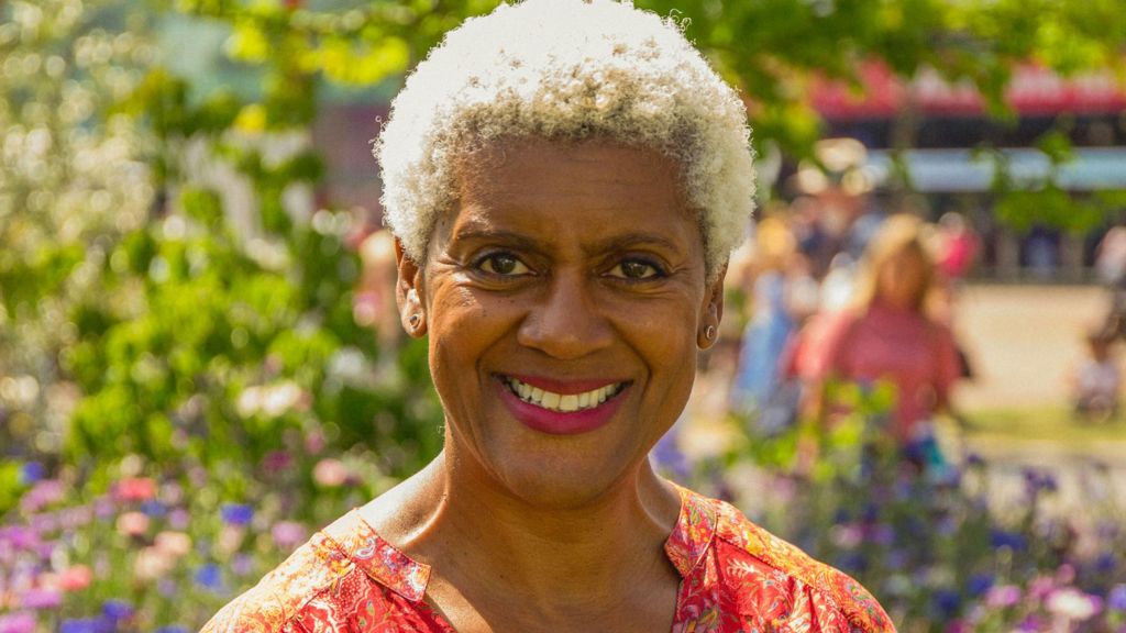 Arit Anderson is smiling and has short bleach blonde hair. She is wearing a colorful dress, and is standing in front of a garden.